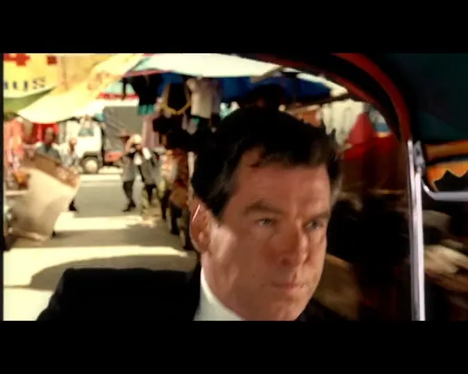 A man in a suit, Pierce Brosnan, looks intently forward while riding in a tuk-tuk through a busy, sunny outdoor market.
