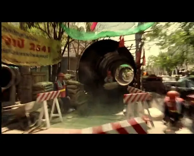 A tuk-tuk speeds through a chaotic construction site, kicking up dust and narrowly avoiding workers and equipment, under a bright sunny sky.
