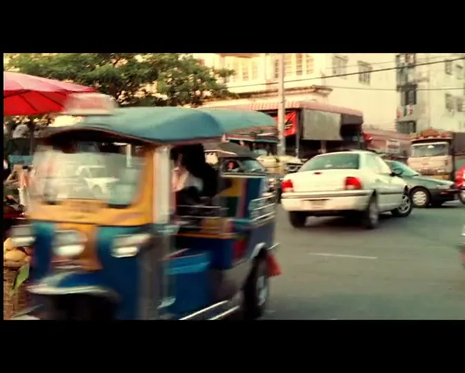 A blue and yellow tuk-tuk drives past a fruit stand on a busy street in an Asian city during the day.