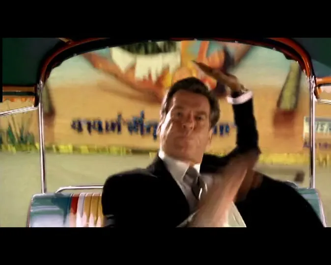 A man in a suit is seen from the chest up, holding onto the roof of a moving tuk-tuk, with a blurred background of a beach scene.