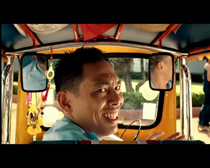 A male tuk-tuk driver, wearing a light blue shirt, turns his head and smiles at the camera while driving his yellow tuk-tuk.