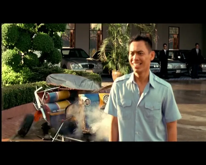 A smiling Asian man in a light blue shirt stands in front of a colorful tuk-tuk that explodes into pieces, emitting smoke, outside a building.