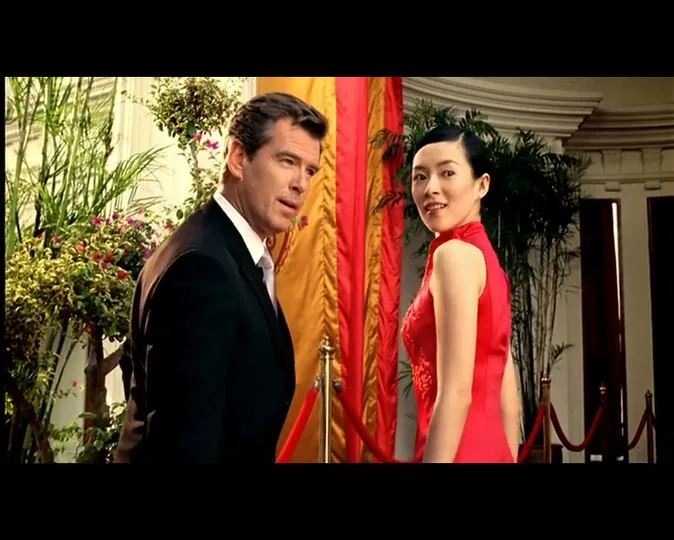 A man in a suit and a woman in a red dress look back at the camera, standing in front of a building with plants and red and gold fabric.
