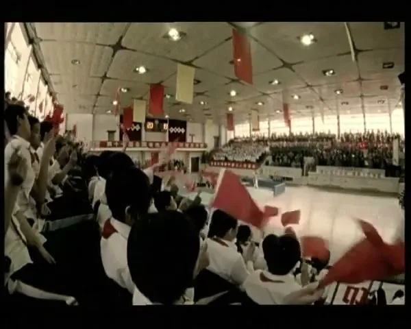 A wide shot of a large crowd of people waving red flags in a stadium, with a scoreboard visible in the background.