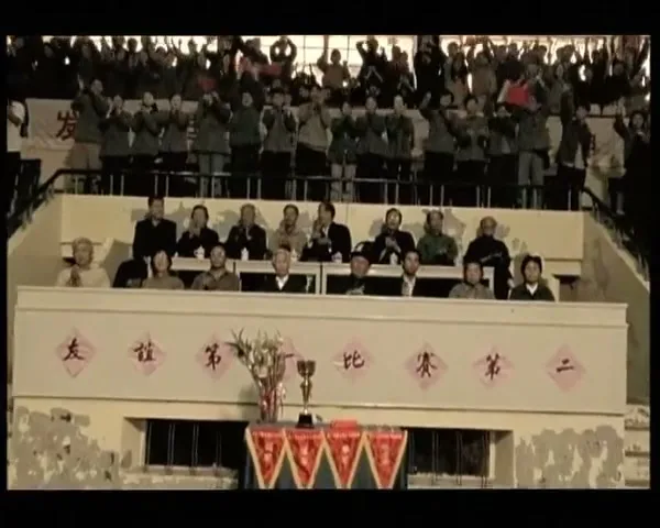A wide shot shows a crowd of people cheering in a stadium, with officials seated below them and a trophy on a table in the foreground.