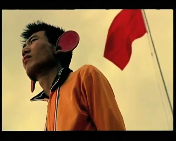 A young man with large, red, paddle-like ears looks up, standing outdoors with a red flag waving in the background.