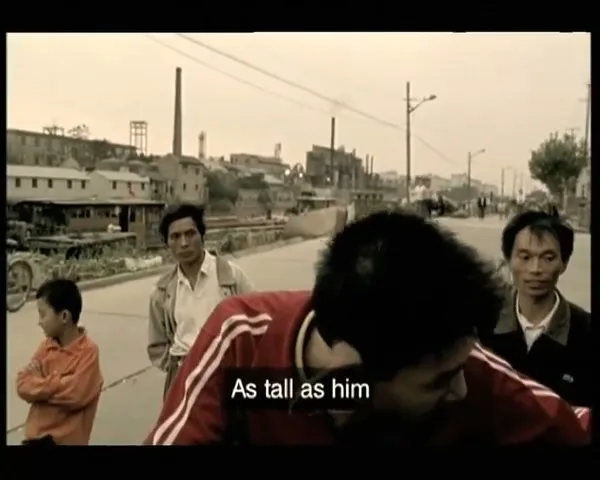 A young man with large red ping-pong paddles attached to his ears talks to the camera on a street, surrounded by onlookers.