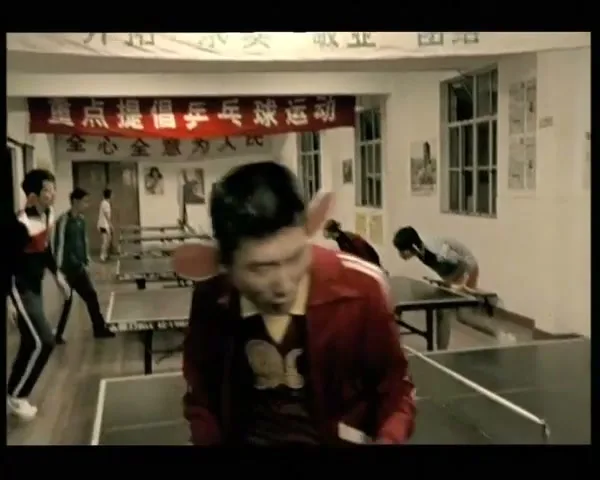 A man with large, prosthetic ears and a red jacket plays ping pong in a busy hall, then runs through a crowded outdoor market.