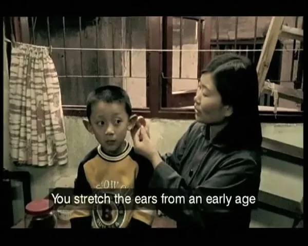 A woman with dark hair in a dark shirt gently pulls on the ear of a young boy in a yellow and black shirt, who looks forward with wide eyes in an interior setting.