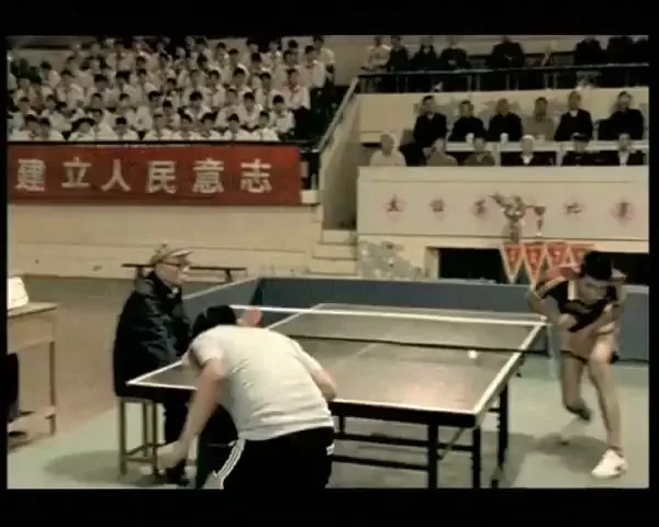 Two male table tennis players compete in a stadium, with one player wearing a red and yellow uniform and the other a grey shirt.