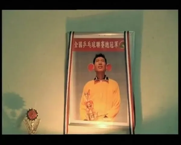 A framed photo of a young man with large, red, cartoonish ears, wearing a yellow shirt and holding a trophy, hangs on a light green wall next to a smaller trophy.
