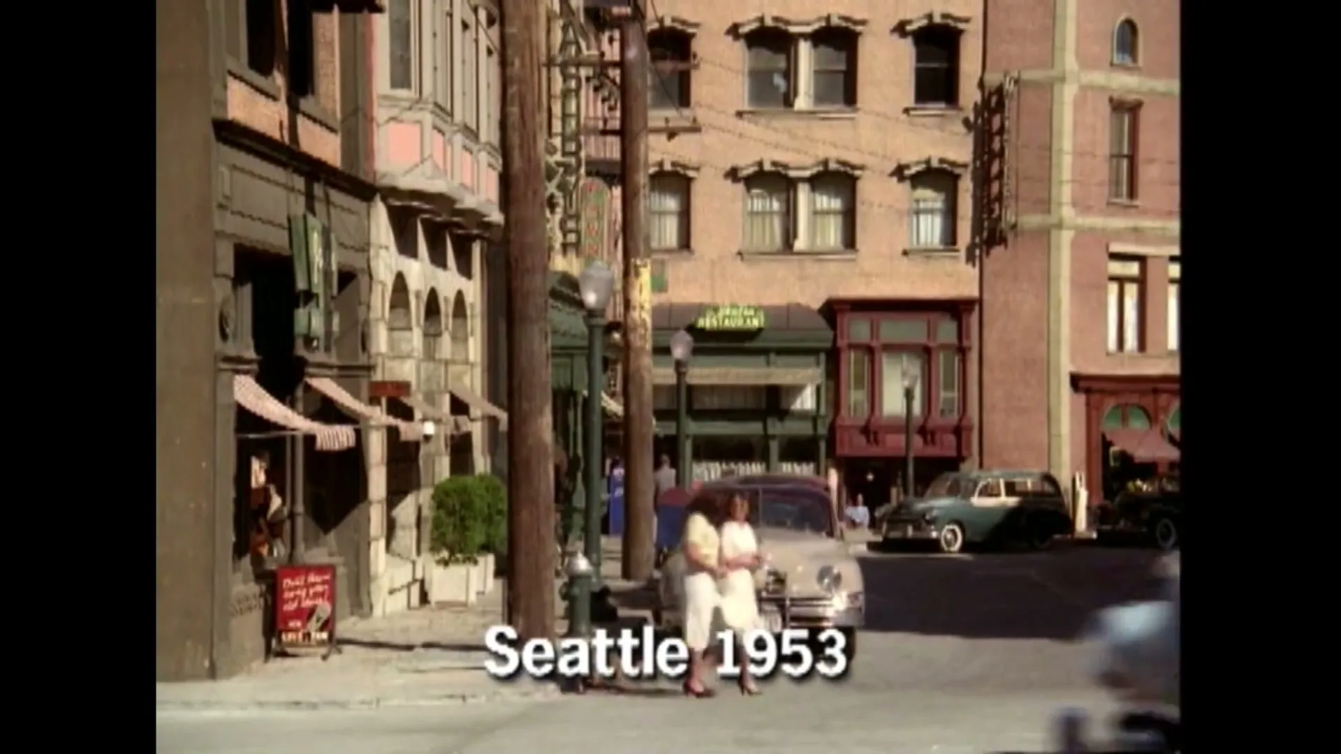 A wide shot of a bustling street in Seattle in 1953, with vintage cars and pedestrians, as a light blue car drives past the camera.