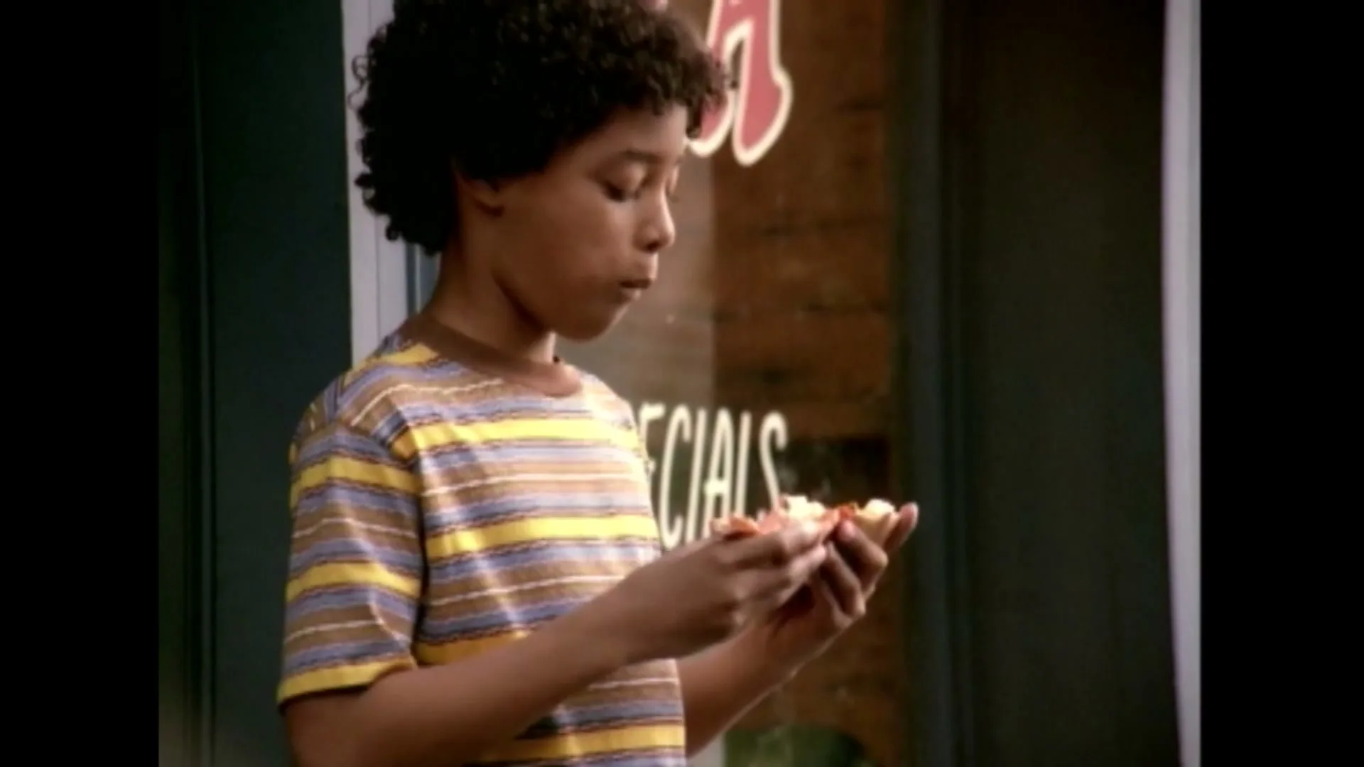 A young boy with curly hair in a striped shirt eats a slice of pizza outside a store.