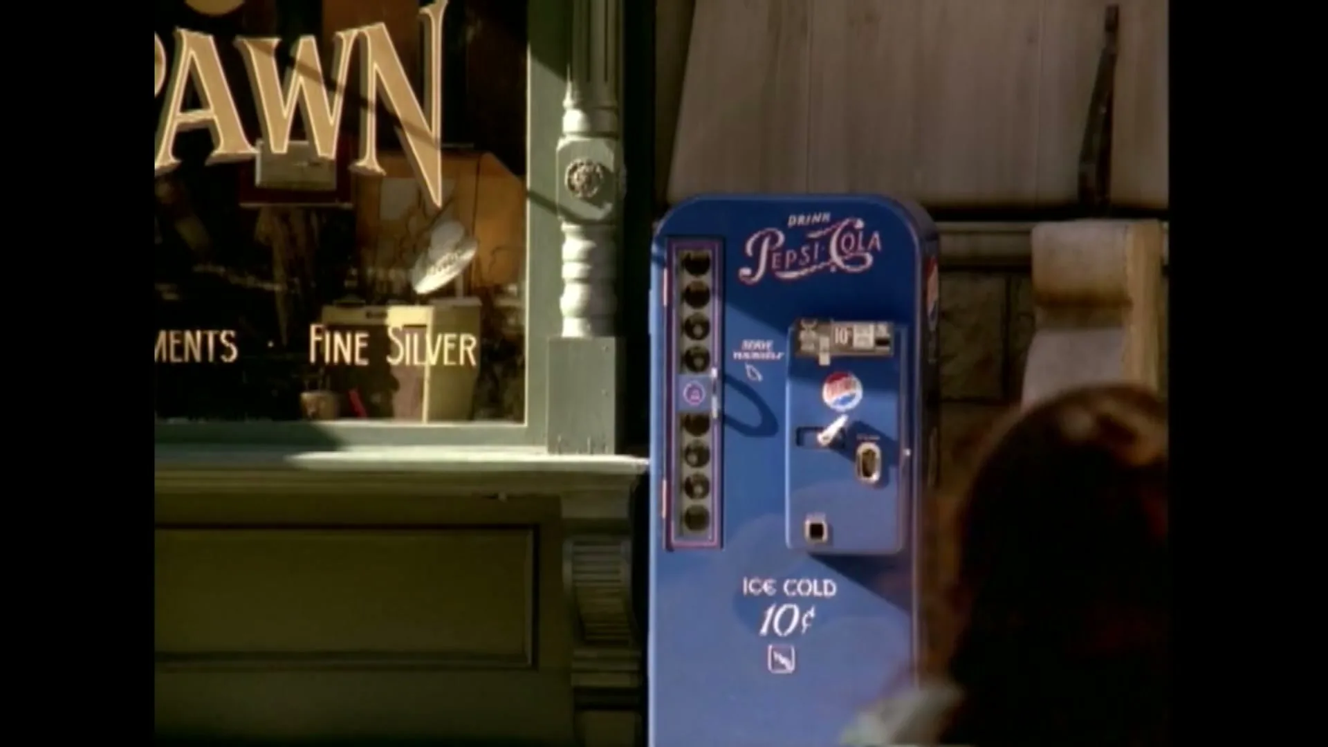A woman with brown hair walks past a blue vintage Pepsi-Cola vending machine on a sunny street next to a pawn shop.