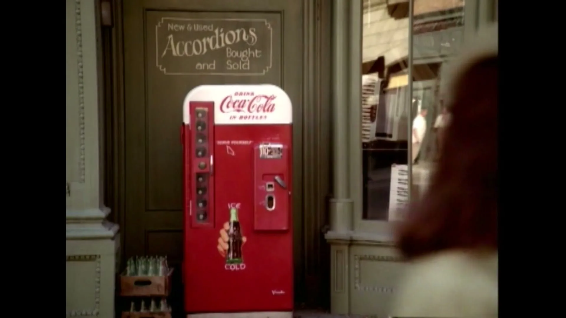 Two young women walk past a vintage Coca-Cola vending machine outside a shop that buys and sells accordions on a sunny day.