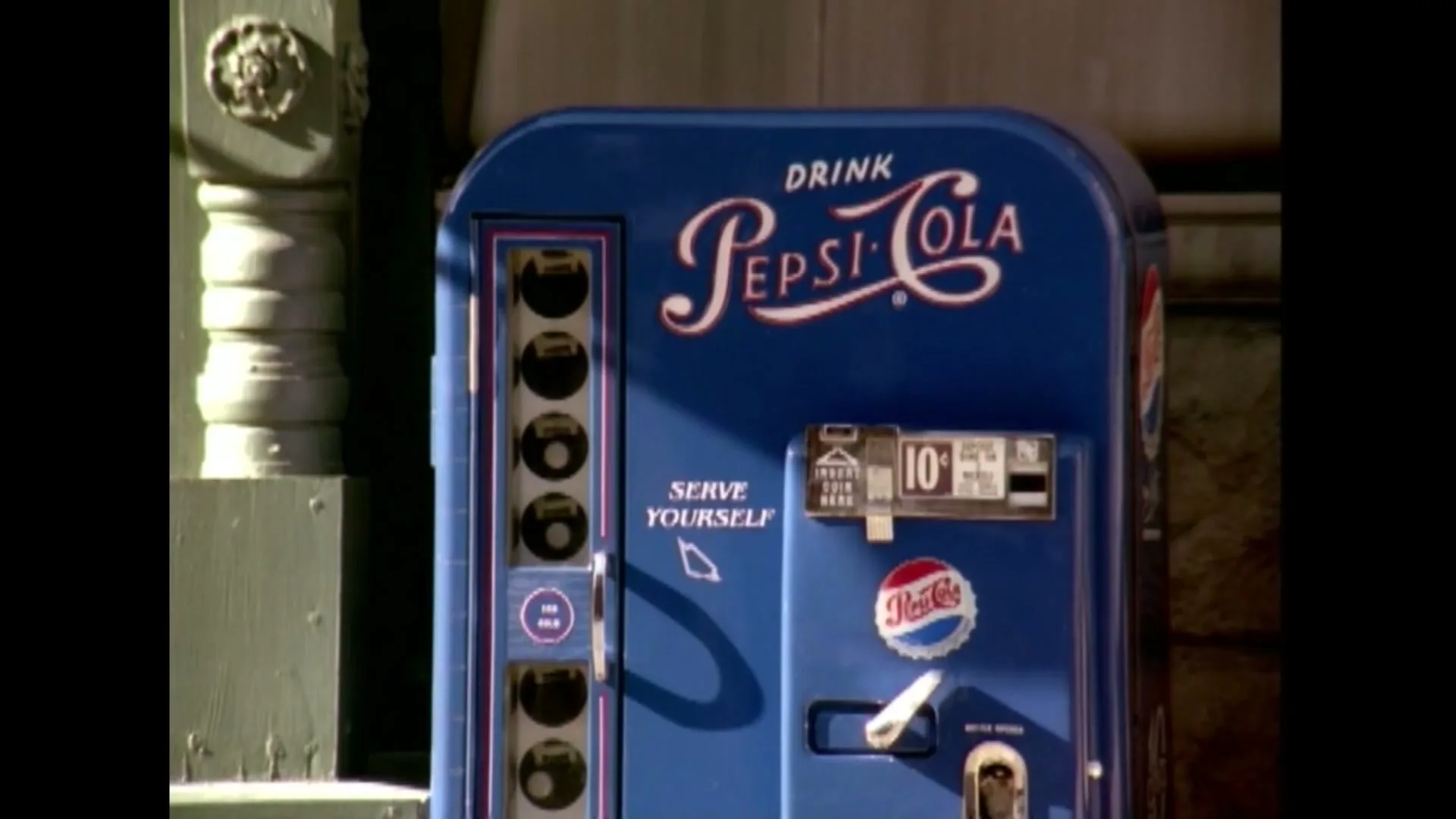 A vintage blue Pepsi-Cola vending machine with a white logo is shown in a medium shot, with a cursor pointing to "SERVE YOURSELF" on the machine.