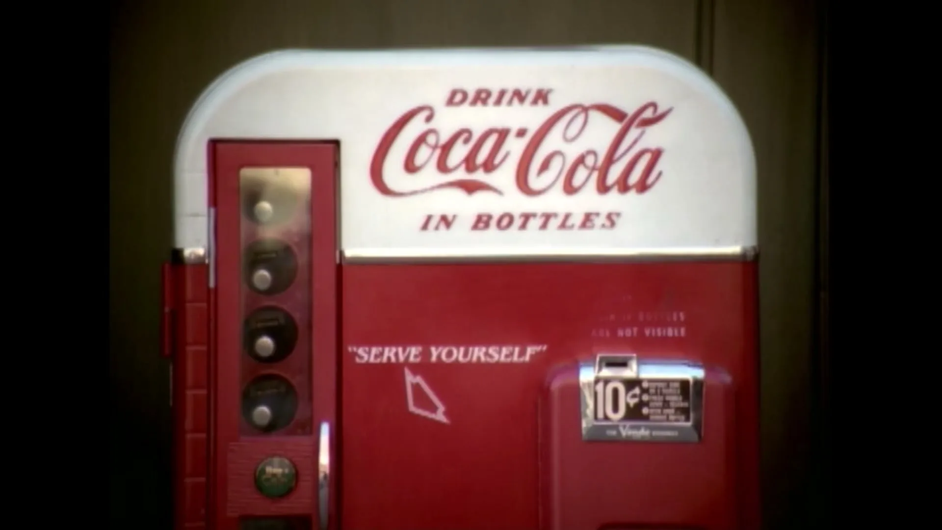 A vintage red and white Coca-Cola vending machine with "Drink Coca-Cola in Bottles" and "Serve Yourself" text is shown in a medium shot.