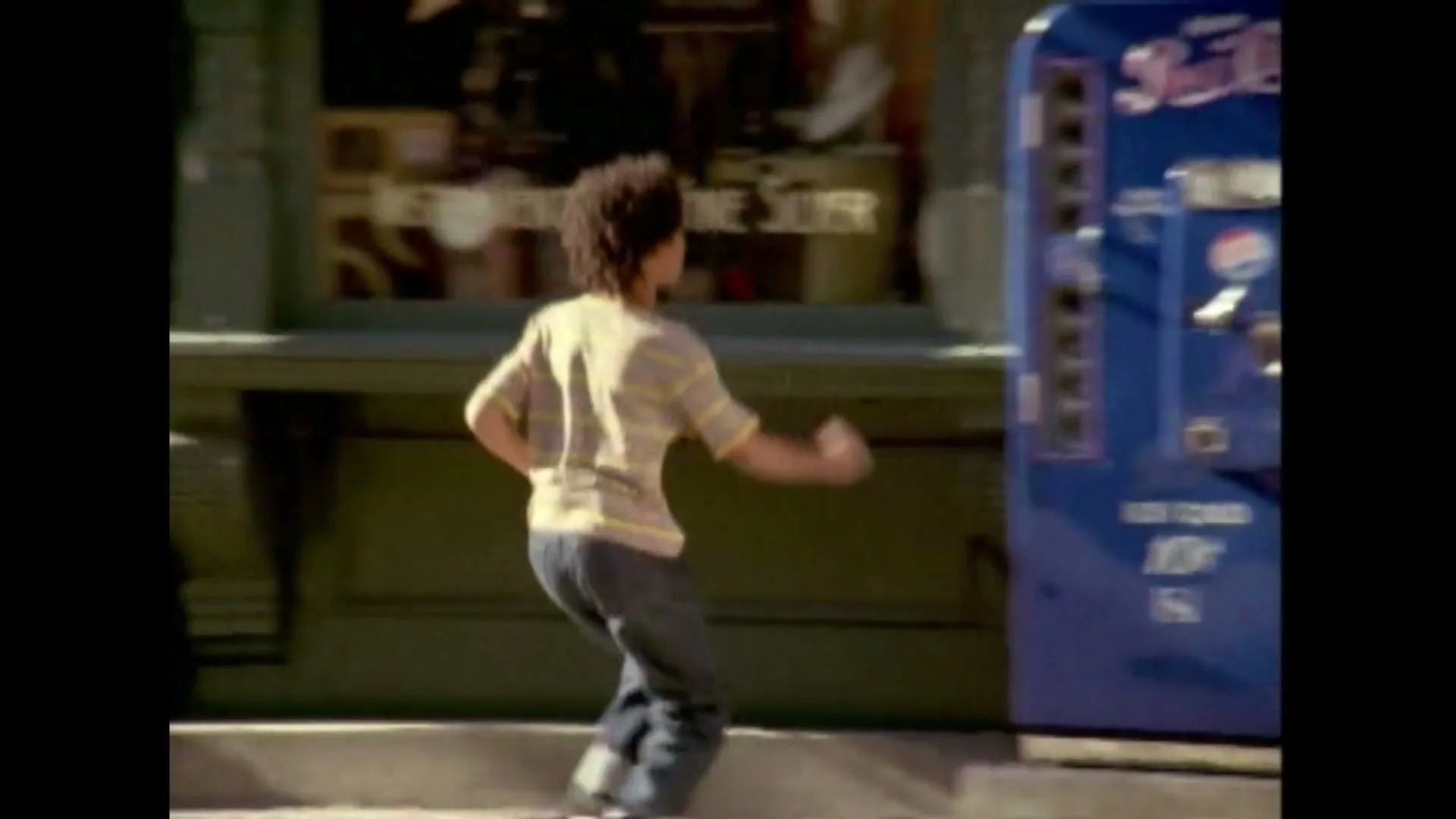 A young boy with curly hair runs past a blue Pepsi vending machine on a sunny street, as a woman walks by.