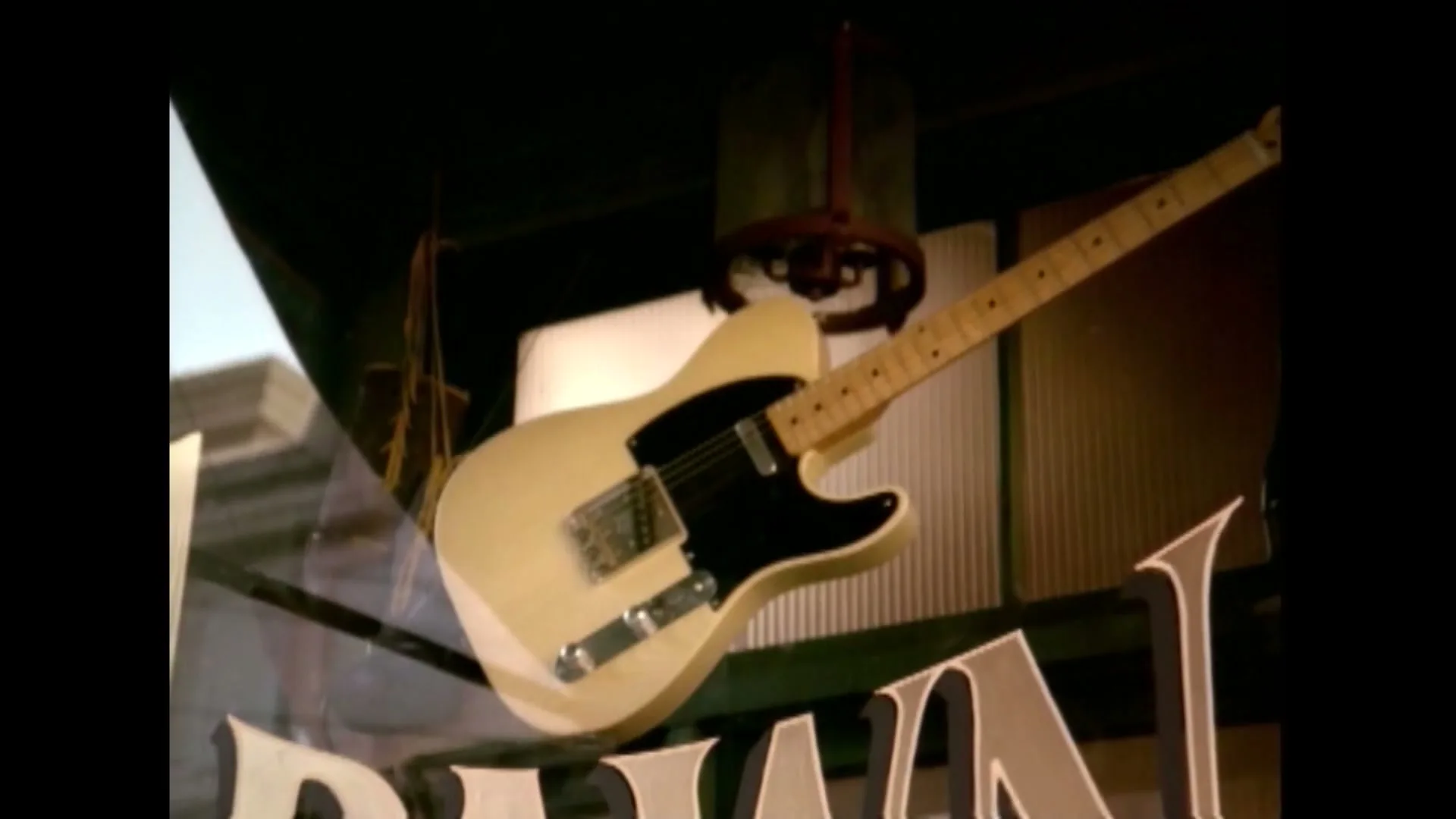 A cream-colored electric guitar with a black pickguard hangs in a store window, with reflections of buildings visible.