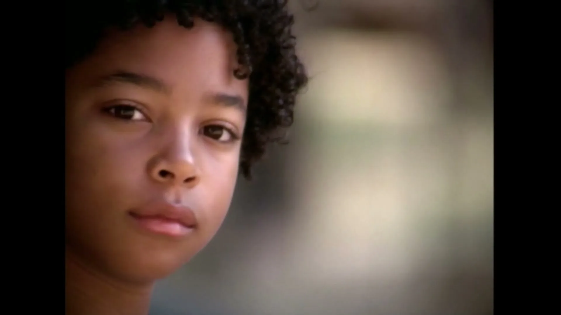 A young Black boy with curly hair looks directly at the camera in a medium closeup shot, outdoors during the day.