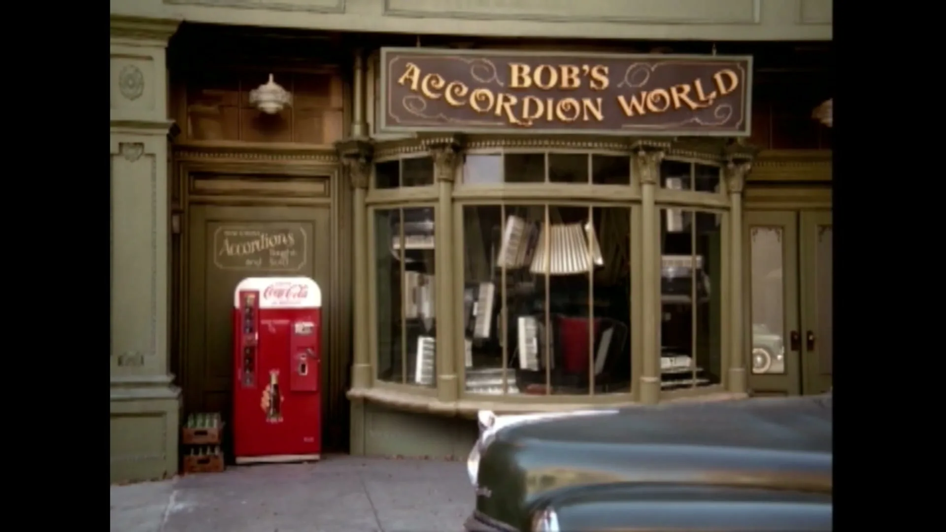 Two men in suits walk past a vintage Coca-Cola vending machine outside "Bob's Accordion World" on a city street during the day.