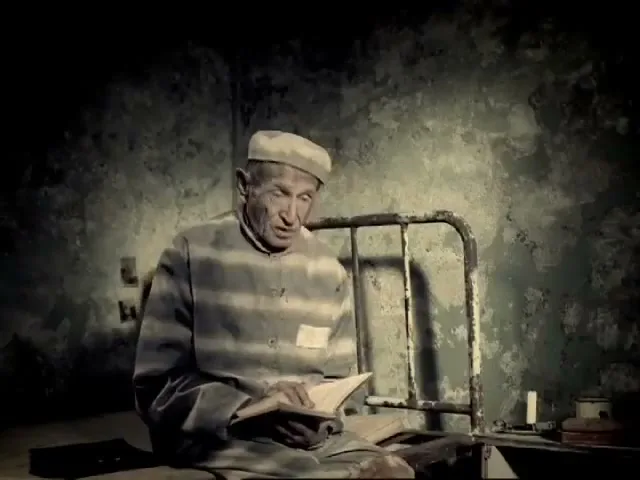 An elderly male prisoner in a striped uniform sits on the floor of his cell, reading a book, then looks up.