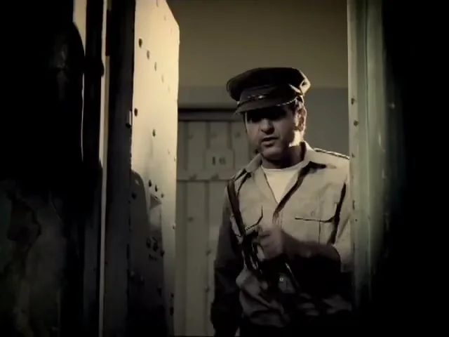 A male prison guard in a uniform opens a cell door, looking sternly into the cell in a dark, gritty prison.