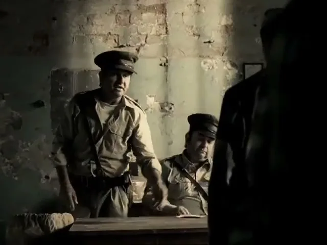 Two male guards in uniform are at a desk in a dimly lit, run-down room, with one guard leaning forward and gesturing.