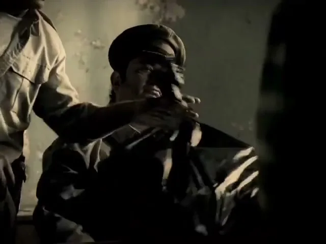 A male prison guard in a uniform sits at a desk, looking sternly as another guard places a black refuse bag on his lap in a dimly lit room.