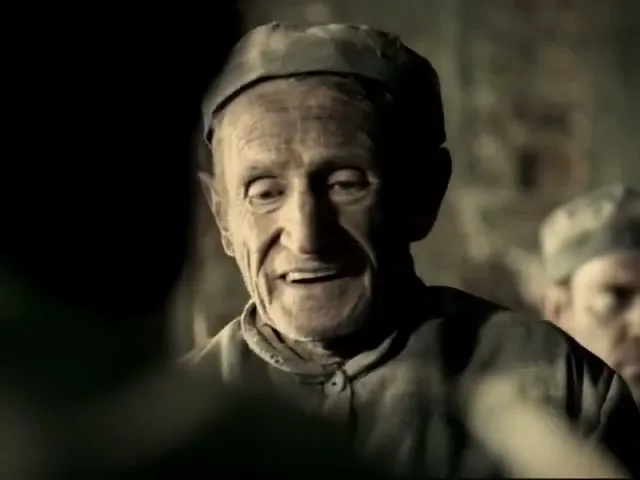 An elderly male prisoner with a dirty face and uniform smiles, looking at someone off-screen in a dimly lit prison.