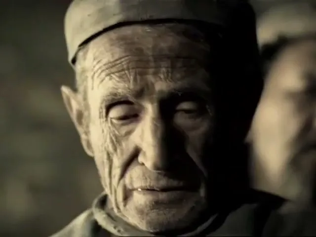 An elderly male prisoner with a cap and dirty clothes looks up with a sad expression in a dark, dirty prison cell.