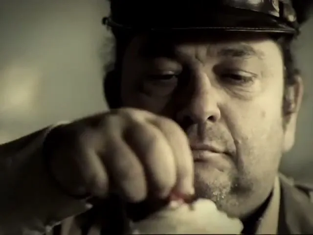 A man in a uniform and hat eats popcorn, looking directly at the camera in a close-up shot.