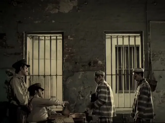 Two prison guards and two prisoners stand in a dimly lit prison cell block, with one prisoner holding a small object.