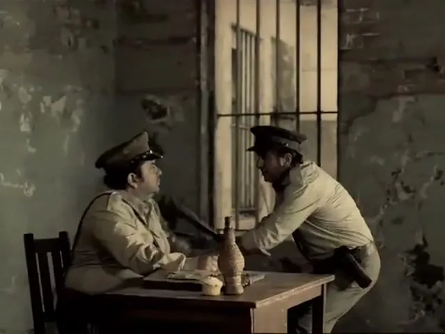 Two male prison guards in uniform are fighting at a desk in a prison cell, with one guard hitting the other with a baton.