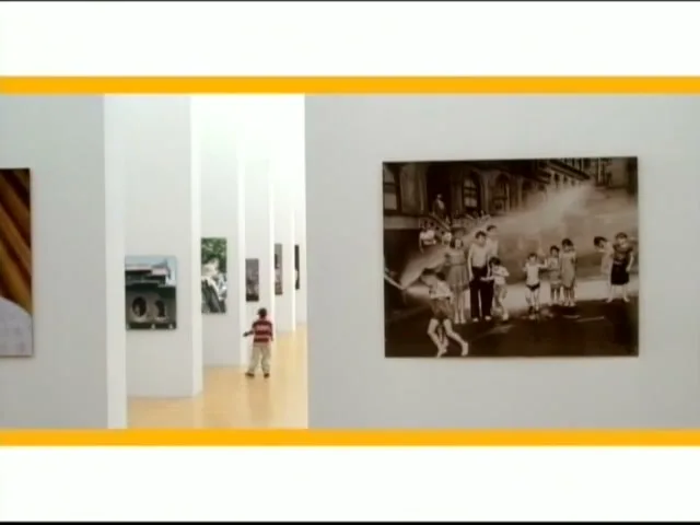A young boy in a striped shirt and khaki pants walks through a modern art gallery with white walls, looking at various framed photographs.