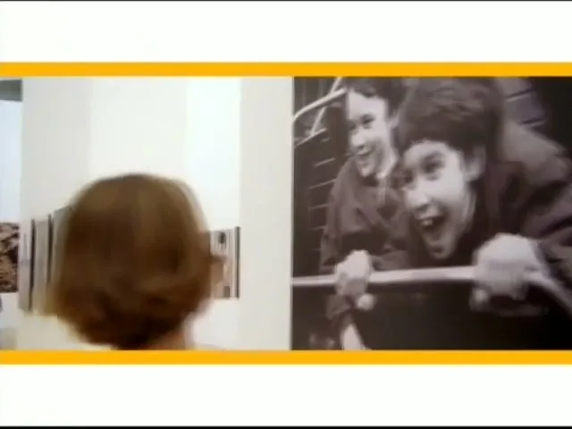 A young woman with short brown hair views a large black and white photograph of two excited children in a brightly lit art gallery.