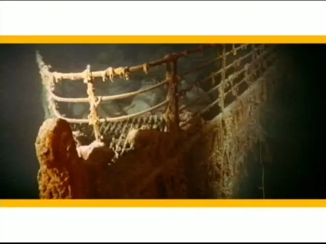 The rusted railing of the sunken Titanic ship is seen underwater, covered in marine growth, with dark water in the background.