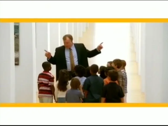 A male teacher in a suit stands in a bright hallway, gesturing to a group of diverse children gathered around him, as if on a field trip.