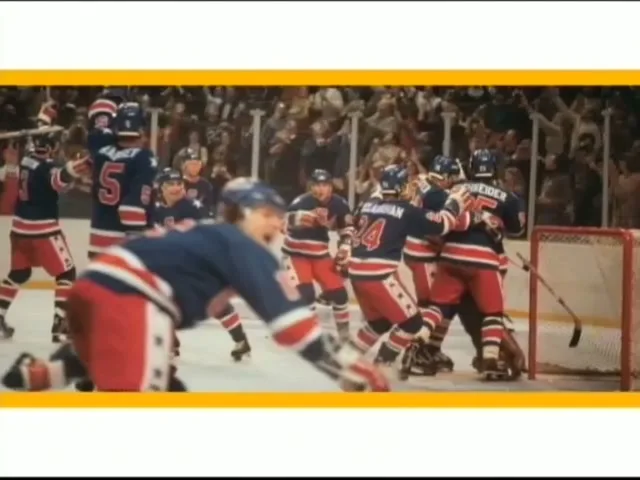 A group of male ice hockey players in blue, red, and white uniforms celebrate a goal on the ice rink, with a crowd in the background.