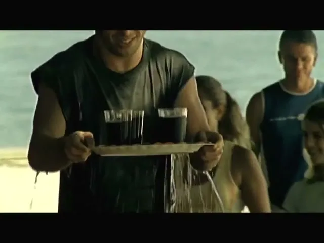 A man in a wet black shirt carries a tray of drinks, smiling as water drips from the tray, with other people and the ocean in the background.