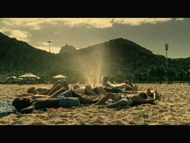 A group of young people lie in a circle on a sandy beach, sunbathing as water sprays upwards from the center of their formation.