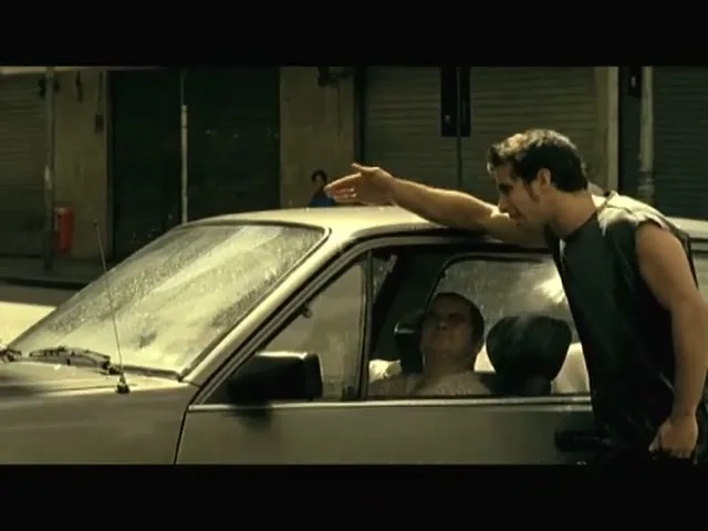 A young man in a black tank top leans into a car window, talking to an older man inside, on a sunny street.