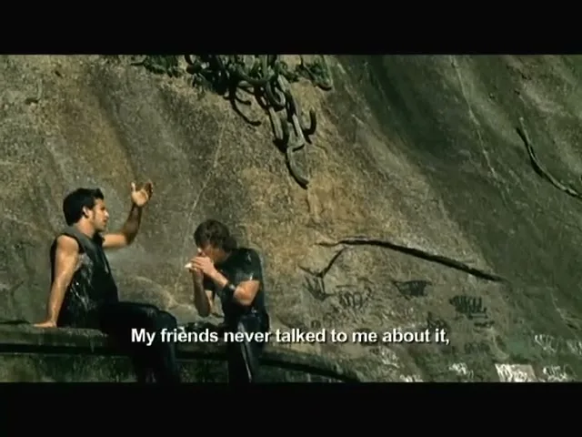 Two young men, wet and in dark clothes, sit on a ledge against a rocky wall with graffiti, one talking and the other applying something to his arm.