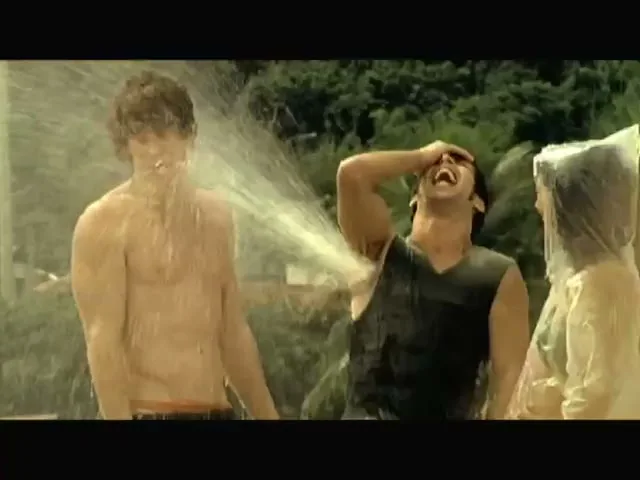 A group of young adults on a beach, two men are being sprayed with water, and a woman in a clear poncho laughs, surrounded by lush green hills.