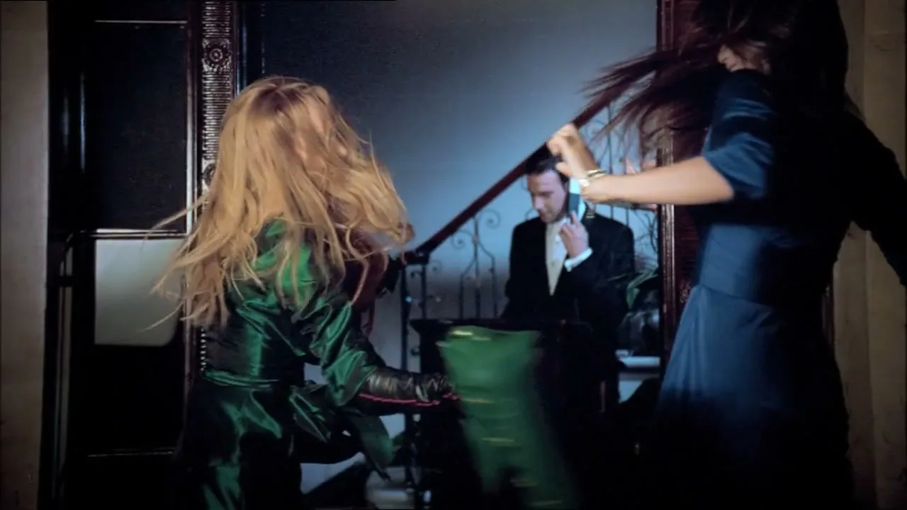 Two women in a hallway fight over a green handbag, while a man in a suit is reflected in a mirror, talking on a phone.
