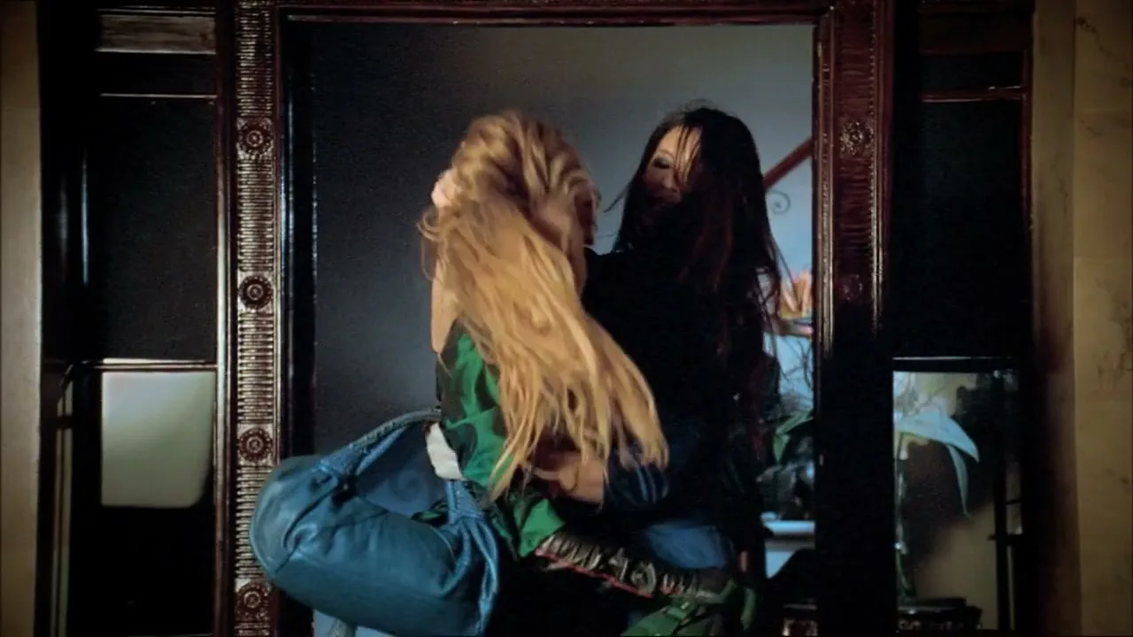 Two women with long hair, one in a green top and the other in a blue top, are playfully fighting over a handbag in a room with a large mirror.