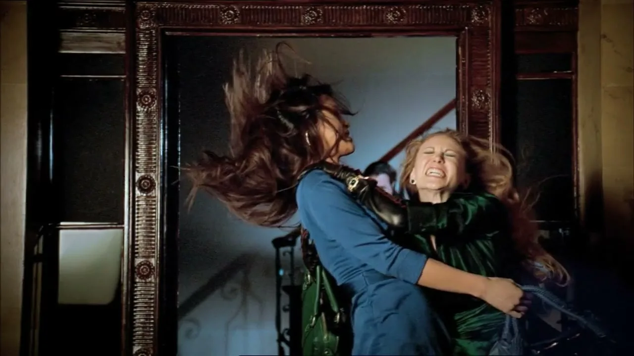 Two women, one in a blue dress and the other in a green top, are fighting over a handbag in a hallway with a large mirror.
