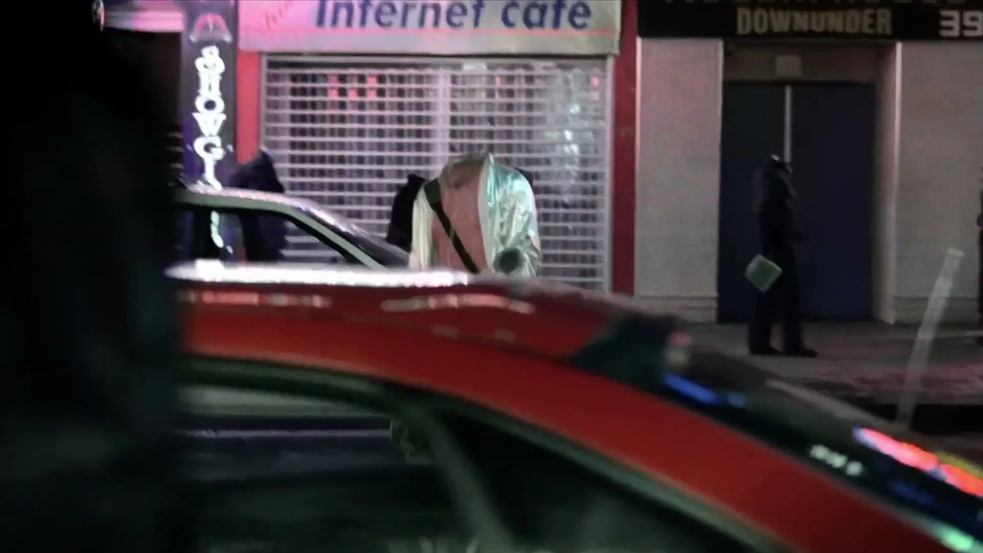 An invisible man in a shiny jacket and pink shirt walks past an internet cafe and a red car on a city street at night.