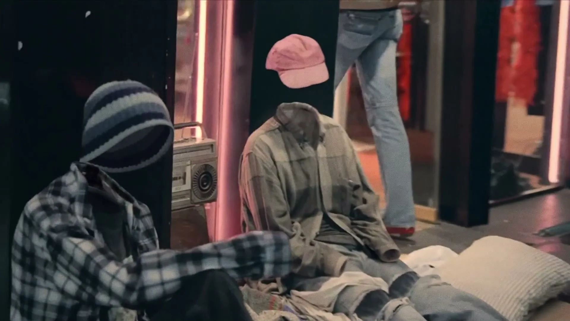 Two sets of clothes, appearing to be worn by invisible people, sit on the ground next to a boombox on a city street at night.