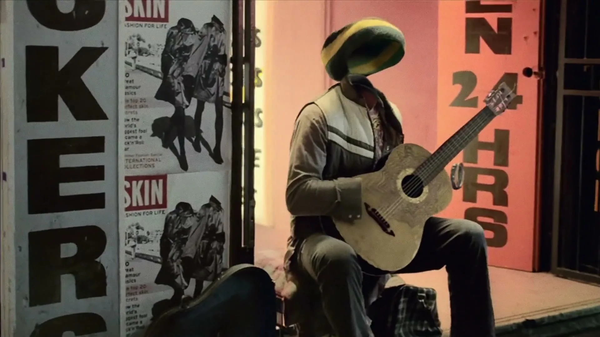An invisible man wearing a rasta hat and jacket sits on a stool, playing an acoustic guitar on a city street at night.